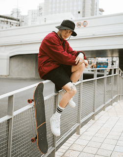 Man sitting on bridge railing with skateboard wearing bucket hat and red shirt