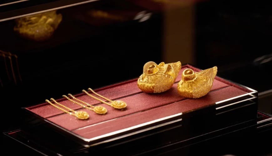 Three Laopu Gold golden necklaces in an illuminated display box, next to two golden ducks